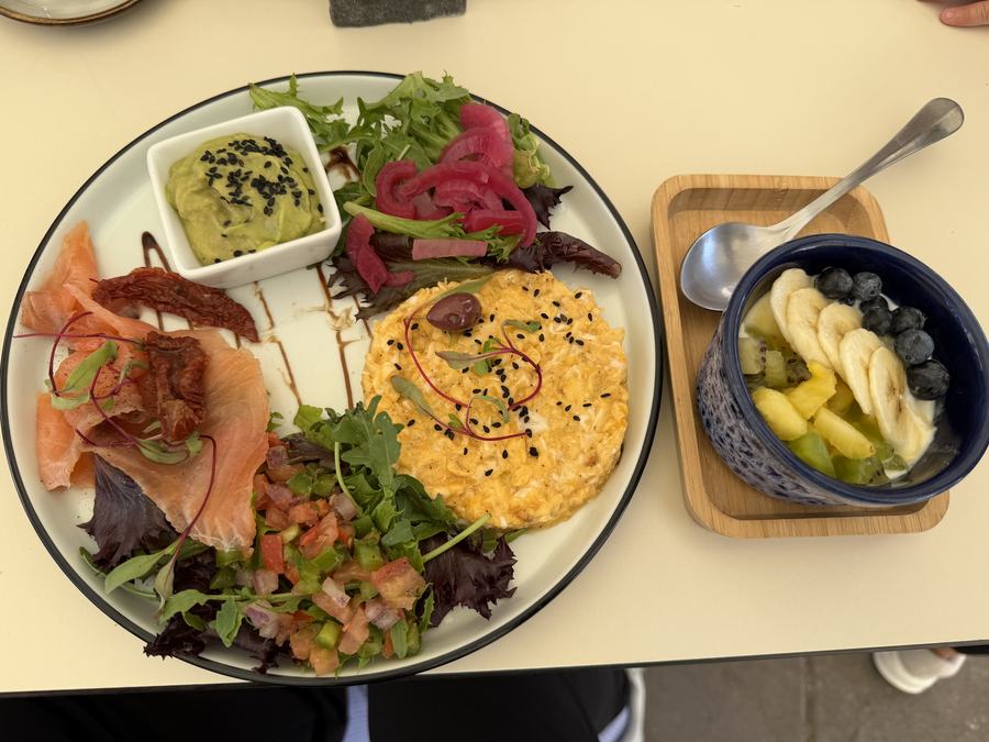 Breakfast plate with scrambled eggs, smoked salmon, greens, tomato salsa, pickled onion and a side bowl of yogurt with fruit.